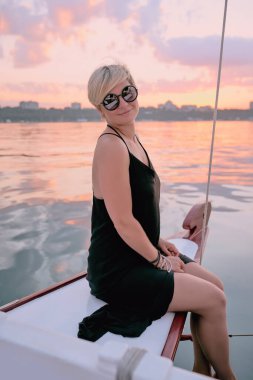 Güneş gözlüklü ve uzun siyah elbiseli güzel sarışın kadın yatta günbatımını seyrediyor. Seyahat, macera, rahatlama, rahatlama konsepti 