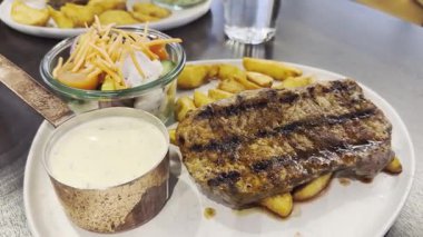 Lokantada servis edilen beyaz tabakta fırında patatesli orta pişmiş biftek. Üst manzara, düz yatış. Orta pişmiş bifteğin yanında kızarmış patates ve kremalı sosla servis ediliyor. Servis edildi.