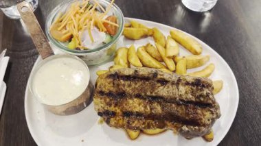 Lokantada servis edilen beyaz tabakta fırında patatesli orta pişmiş biftek. Üst manzara, düz yatış. Orta pişmiş bifteğin yanında kızarmış patates ve kremalı sosla servis ediliyor. Servis edildi.