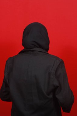 Sırtı kameraya dönük tesettürlü bir kadın, kırmızı arka plan.