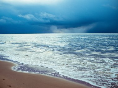 Denizin küçük dalgaları kumla savaşıyor. Deniz dalgaları kumlu sahili temizler. Gökyüzünde yağmur bulutları görünür.