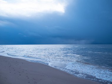 Denizin küçük dalgaları kumla savaşıyor. Deniz dalgaları kumlu sahili temizler. Gökyüzünde yağmur bulutları görünür.