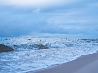 Denizin küçük dalgaları kumla savaşıyor. Deniz dalgaları kumlu sahili temizler. Gökyüzünde yağmur bulutları görünür.