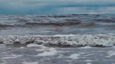 Denizin küçük dalgaları kumla savaşıyor. Deniz dalgaları kumlu sahili temizler. Gökyüzünde yağmur bulutları görünür.