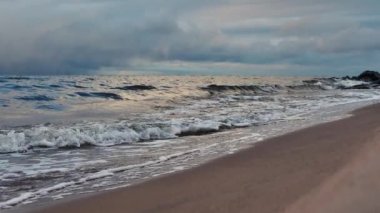 Denizin küçük dalgaları kumla savaşıyor. Deniz dalgaları kumlu sahili temizler. Gökyüzünde yağmur bulutları görünür.
