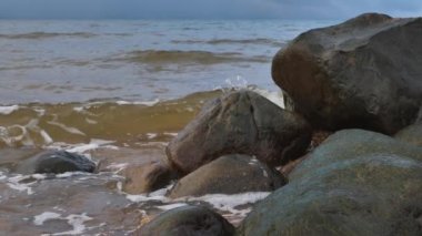 Deniz dalgaları büyük taşları temizler. Gökyüzünde yağmur bulutları görünür.