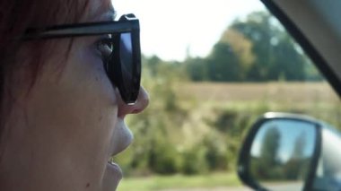 Güneş gözlüklü bir kadın arabanın direksiyonunda konuşuyor.