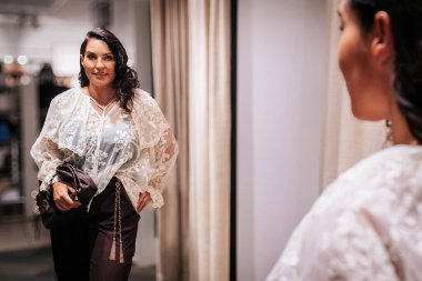 Beyaz çiçekli bluz, altın zincirli siyah pantolon ve soyunma odasında erik çantasıyla yansımasına hayran olan bir kadın..