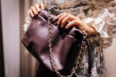 Elinde altın tokalı küçük bir deri çanta tutan bir kadının yakın çekimi. Hafif aydınlatılmış bir soyunma odasında siyah desenli bir elbiseyle eşleştirilmiş..