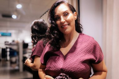 Kalıplı bordo bir elbise içinde zarif bir kadın, altın zincirli bir çanta tutuyor, şık bir ortamda yumuşak krema perdelerine karşı güvenle poz veriyor..