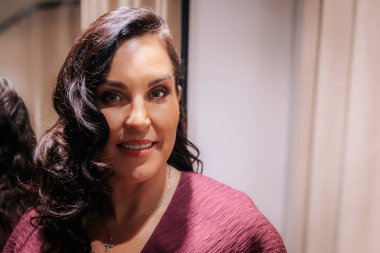 Kalıplı bordo bir elbise içinde zarif bir kadın, altın zincirli bir çanta tutuyor, şık bir ortamda yumuşak krema perdelerine karşı güvenle poz veriyor..