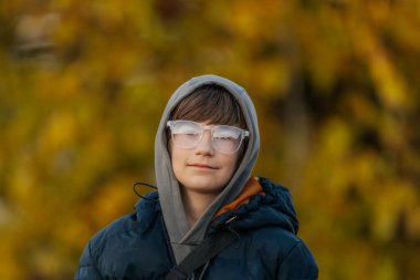 Gri kapüşonlu ve koyu mavi kabarık ceketli çocuk arka planda altın sonbahar yapraklarıyla dışarıda duruyor..
