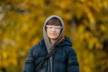 Gri kapüşonlu ve koyu mavi kabarık ceketli çocuk arka planda altın sonbahar yapraklarıyla dışarıda duruyor..