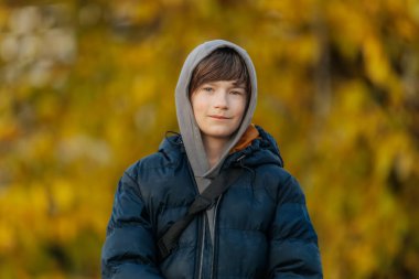 Mavi kabarık ceketli ve gri kapüşonlu gülümseyen çocuk altın sonbahar yapraklarının önünde dikiliyor..