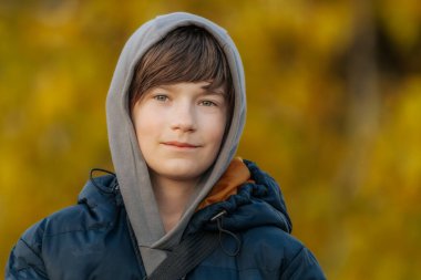 Mavi kabarık ceketli ve gri kapüşonlu gülümseyen çocuk altın sonbahar yapraklarının önünde dikiliyor..