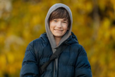 Mavi kabarık ceketli ve gri kapüşonlu gülümseyen çocuk altın sonbahar yapraklarının önünde dikiliyor..