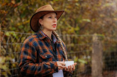 Kahverengi kovboy şapkalı ve ekose ceketli bir kadın sonbaharda dışarıda duruyor, örgülü ve cesur küpeleri kırsal ve taşralı görünümünü tamamlıyor..