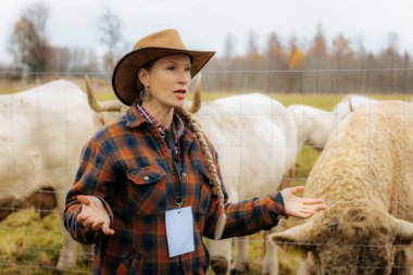 Ekose ceketli ve kovboy şapkalı kadın bulutlu bir günde tel örgülerin arkasında beyaz sığırların yanında duruyor, konuşuyor ya da gözlemliyor. Görünür logo yok.