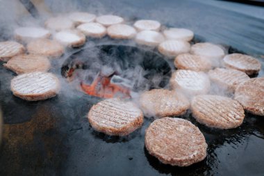Hamburger köfteleri parlayan kömürün üstünde çatal ve spatula kullanarak mangalda cızırdıyor. Buhar ve ızgara izleri görünür..