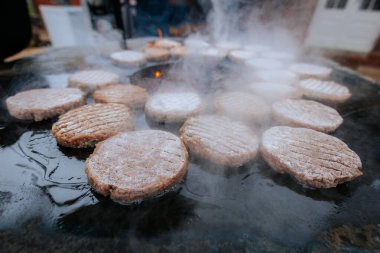 Hamburger köfteleri parlayan kömürün üstünde çatal ve spatula kullanarak mangalda cızırdıyor. Buhar ve ızgara izleri görünür..