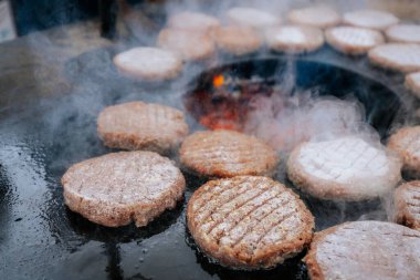 Hamburger köfteleri parlayan kömürün üstünde çatal ve spatula kullanarak mangalda cızırdıyor. Buhar ve ızgara izleri görünür..
