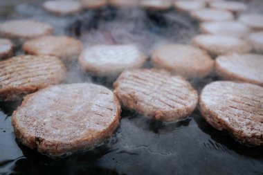 Hamburger köfteleri parlayan kömürün üstünde çatal ve spatula kullanarak mangalda cızırdıyor. Buhar ve ızgara izleri görünür..