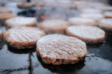 Hamburger köfteleri parlayan kömürün üstünde çatal ve spatula kullanarak mangalda cızırdıyor. Buhar ve ızgara izleri görünür..