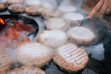 Hamburger köfteleri parlayan kömürün üstünde çatal ve spatula kullanarak mangalda cızırdıyor. Buhar ve ızgara izleri görünür..