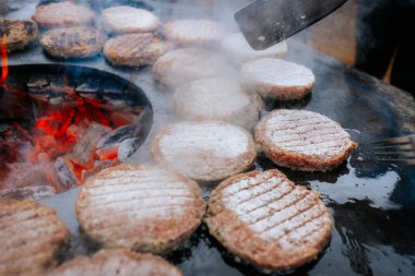 Hamburger köfteleri parlayan kömürün üstünde çatal ve spatula kullanarak mangalda cızırdıyor. Buhar ve ızgara izleri görünür..