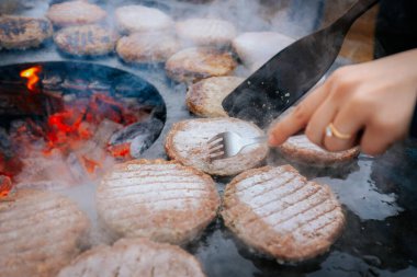 Hamburger köfteleri parlayan kömürün üstünde çatal ve spatula kullanarak mangalda cızırdıyor. Buhar ve ızgara izleri görünür..