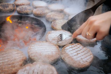 Hamburger köfteleri parlayan kömürün üstünde çatal ve spatula kullanarak mangalda cızırdıyor. Buhar ve ızgara izleri görünür..
