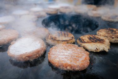 Hamburger köfteleri parlayan kömürün üstünde çatal ve spatula kullanarak mangalda cızırdıyor. Buhar ve ızgara izleri görünür..
