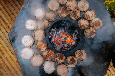 Hamburger köfteleri parlayan kömürün üstünde çatal ve spatula kullanarak mangalda cızırdıyor. Buhar ve ızgara izleri görünür..
