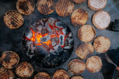 Hamburger köfteleri parlayan kömürün üstünde çatal ve spatula kullanarak mangalda cızırdıyor. Buhar ve ızgara izleri görünür..