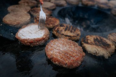 Hamburger köfteleri parlayan kömürün üstünde çatal ve spatula kullanarak mangalda cızırdıyor. Buhar ve ızgara izleri görünür..