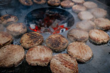 Hamburger köfteleri parlayan kömürün üstünde çatal ve spatula kullanarak mangalda cızırdıyor. Buhar ve ızgara izleri görünür..