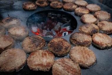 Hamburger köfteleri parlayan kömürün üstünde çatal ve spatula kullanarak mangalda cızırdıyor. Buhar ve ızgara izleri görünür..
