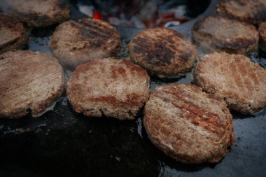 Hamburger köfteleri parlayan kömürün üstünde çatal ve spatula kullanarak mangalda cızırdıyor. Buhar ve ızgara izleri görünür..