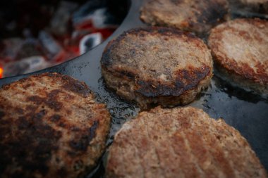Hamburger köfteleri parlayan kömürün üstünde çatal ve spatula kullanarak mangalda cızırdıyor. Buhar ve ızgara izleri görünür..