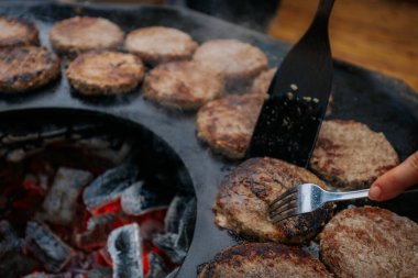 Hamburger köfteleri parlayan kömürün üstünde çatal ve spatula kullanarak mangalda cızırdıyor. Buhar ve ızgara izleri görünür..