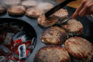 Hamburger köfteleri parlayan kömürün üstünde çatal ve spatula kullanarak mangalda cızırdıyor. Buhar ve ızgara izleri görünür..