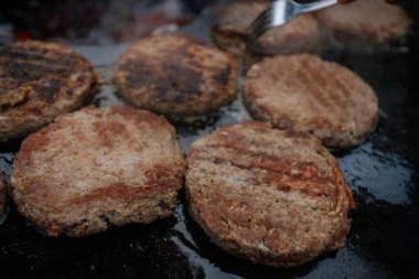 Hamburger köfteleri parlayan kömürün üstünde çatal ve spatula kullanarak mangalda cızırdıyor. Buhar ve ızgara izleri görünür..