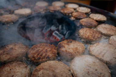 Hamburger köfteleri parlayan kömürün üstünde çatal ve spatula kullanarak mangalda cızırdıyor. Buhar ve ızgara izleri görünür..