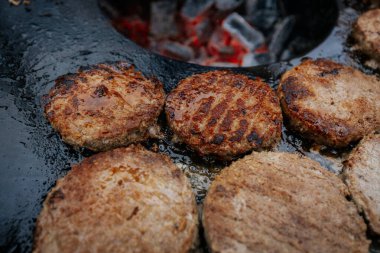 Hamburger köfteleri parlayan kömürün üstünde çatal ve spatula kullanarak mangalda cızırdıyor. Buhar ve ızgara izleri görünür..