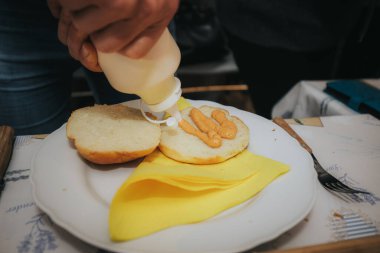 Salatalı, çedarlı, soğanlı, sulu bir cheeseburgerin yakın çekimi, ve altın bir çöreğin içinde bir biftek köftesi, canlı bir iç mekanda beyaz bir tabakta servis edilir..