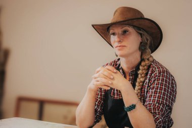 Kahverengi kovboy şapkası takan ve kareli kırmızı ekose gömlekli bir kadın ellerini sıkarak yavaşça gülümseyerek bir masada oturuyor. Uzun sarı örgüsü ve süslü dairesel küpeleri ona köylü havası veriyor..