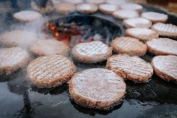 Hamburger köfteleri parlayan kömürün üstünde çatal ve spatula kullanarak mangalda cızırdıyor. Buhar ve ızgara izleri görünür..
