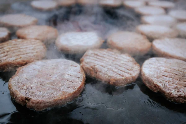 Hamburger köfteleri parlayan kömürün üstünde çatal ve spatula kullanarak mangalda cızırdıyor. Buhar ve ızgara izleri görünür..