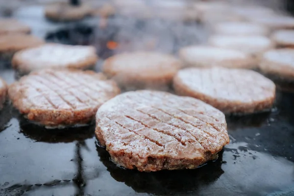 Hamburger köfteleri parlayan kömürün üstünde çatal ve spatula kullanarak mangalda cızırdıyor. Buhar ve ızgara izleri görünür..
