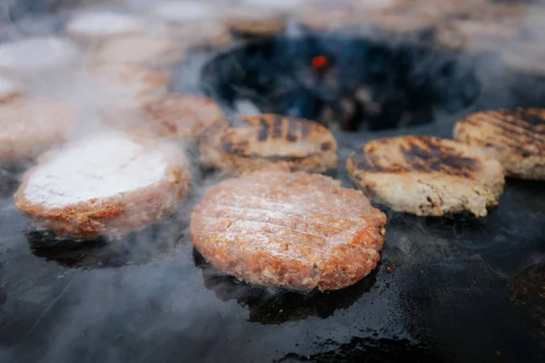 Hamburger köfteleri parlayan kömürün üstünde çatal ve spatula kullanarak mangalda cızırdıyor. Buhar ve ızgara izleri görünür..